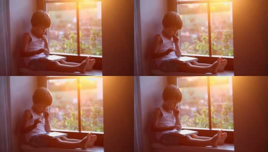 可爱的小男孩，玩着平板电脑，坐在窗罩上，夕阳落在窗后高清在线视频素材下载