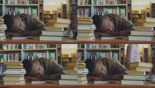 非裔美籍英俊年轻男学生正坐在图书馆里堆满书的桌子旁。他的头躺在桌子上，他正在睡觉，很累。高清在线视频素材下载