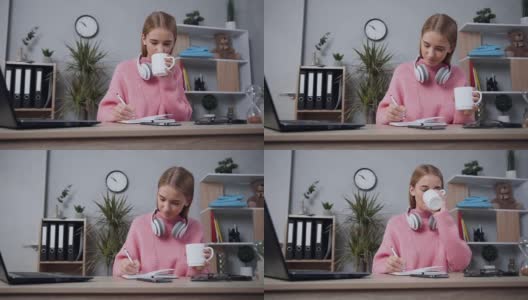满意的微笑迷人的现代女孩在粉红色的毛衣喝茶和写东西到笔记本的特写高清在线视频素材下载