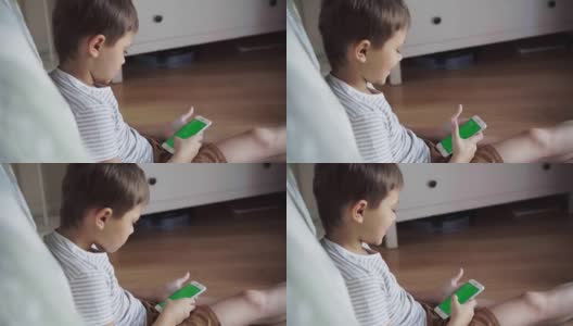 小男孩坐在地板上看手机上的内容。绿屏模型。带着智能手机的孩子的后视图高清在线视频素材下载