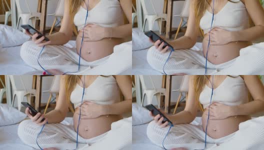 一个年轻的孕妇坐在床上，耳机里戴着智能手机。她积极地在网上交流——通过她的智能手机写信和发送信息高清在线视频素材下载