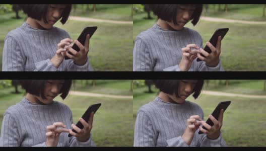 一个亚洲女孩正在室外的绿色公园里看智能手机。4k镜头慢动作高清在线视频素材下载