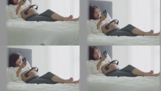 年轻的亚洲孕妇在家里躺在卧室的床上，为未出生的孩子打开音乐，使用智能手机。高清在线视频素材下载