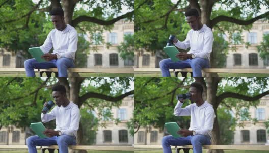 一名自信、专注的非洲裔美国人坐在大学院子的长凳上，喝着早茶，用平板电脑上网。集中的聪明英俊的学生在网上学习的肖像。高清在线视频素材下载