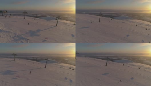 在阳光明媚的冬季日落，人们乘坐缆车在滑雪坡上滑雪和滑板。鸟瞰图。无人机正在横向飞行。远景。高清在线视频素材下载