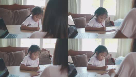 亚洲女童学生做她的家庭作业，而母亲在家工作。由于Covid - 19大流行，在隔离期间，孩子在家上学，父母用电脑在家工作。高清在线视频素材下载