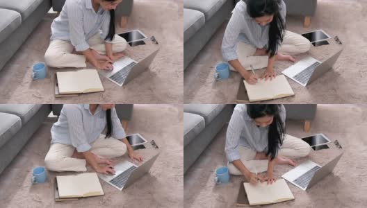 一位年轻的亚洲女商人正在笔记本电脑上寻找信息，她在笔记本上做笔记，她坐在家里客厅的地毯上。商业女性和工作理念。高清在线视频素材下载