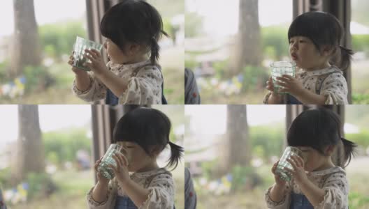 在一家餐馆里，一个天真的亚洲小女孩举起吐司，从窗边的杯子里喝水，做出滑稽的表情高清在线视频素材下载