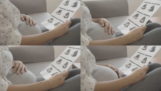 亚洲孕妇看超声波图像和触摸她的腹部坐在沙发上。高清在线视频素材下载