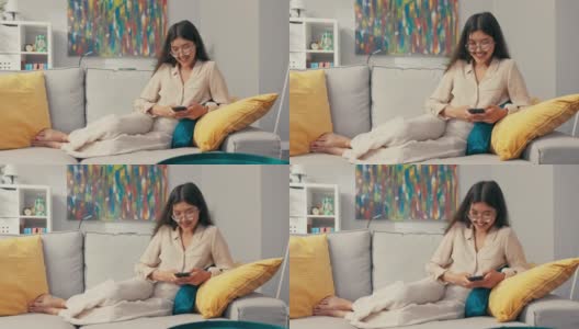 一个有着独特异国风情的美丽女人坐在房间的沙发上，赤脚摊在床上，用手指敲着电话，给朋友发短信，大笑着，微笑着高清在线视频素材下载