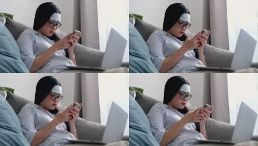 年轻的亚洲女商人感到头痛和发烧。她在家里客厅的沙发上用智能手机和笔记本电脑工作。在家工作，商业女性和工作理念。高清在线视频素材下载