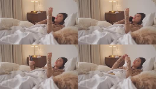 一名亚洲华裔妇女躺在床上，盖着毯子，患感冒和流感，用智能手机和朋友聊天，用数字体温计测量体温高清在线视频素材下载