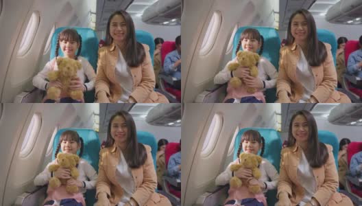 亚洲母女坐在飞机上靠近窗口，微笑着看着镜头。当飞机在天空中飞行时，女孩抱着熊娃娃。航空运输的概念。高清在线视频素材下载