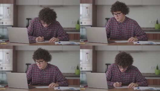 卷发男学生英俊的年轻男孩戴着眼镜正在家里用笔记本电脑学习打字，在笔记本上写字。大学生使用笔记本电脑观看远程在线学习研讨会高清在线视频素材下载