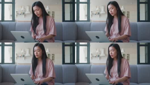 年轻沉思的亚裔女性坐在客厅的沙发上，在笔记本电脑上工作，打字，环顾四周，看起来心烦意乱，失望和沮丧高清在线视频素材下载