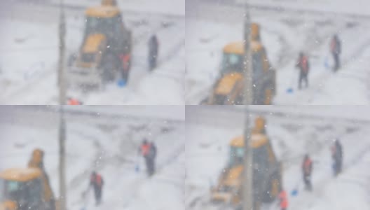 在冬天的暴风雪中清扫街道。工人们拿着铁锹和带着铲斗的拖拉机在离焦的道路上清理积雪。选择性聚焦，浅景深。大雪高清在线视频素材下载
