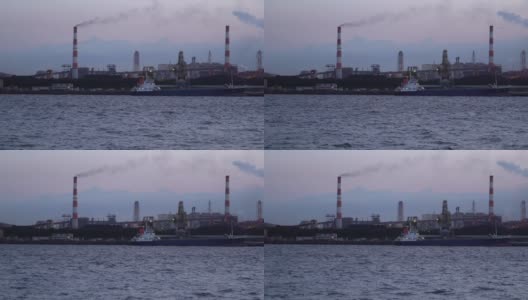 一艘工业船在燃煤电厂前的码头卸煤，烟囱里冒出滚滚浓烟。高清在线视频素材下载