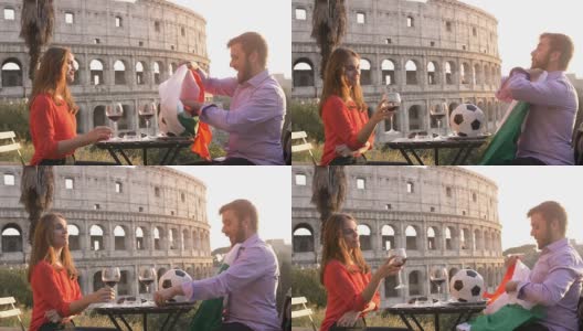 日落时分，在罗马罗马圆形大剧场前，一对浪漫的年轻情侣坐在餐厅的餐桌前，一个年轻人和他的女友正在用意大利国旗踢足球高清在线视频素材下载