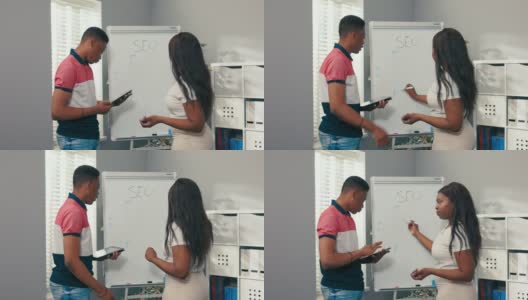 一个非洲裔美国女孩站在办公室的白板前，拿着马克笔，画一个图表，分析它，工作在一个同事检查平板电脑上的图表的监督下进行高清在线视频素材下载