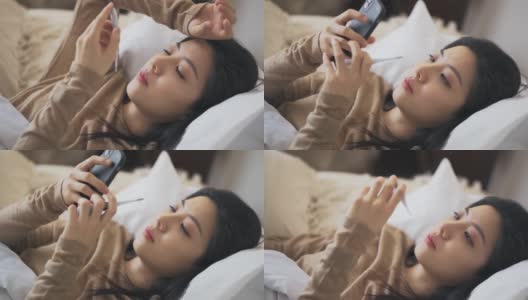 一名亚洲华裔妇女躺在床上，盖着毯子，患感冒和流感，用智能手机给朋友发短信，用数字温度计测量体温高清在线视频素材下载