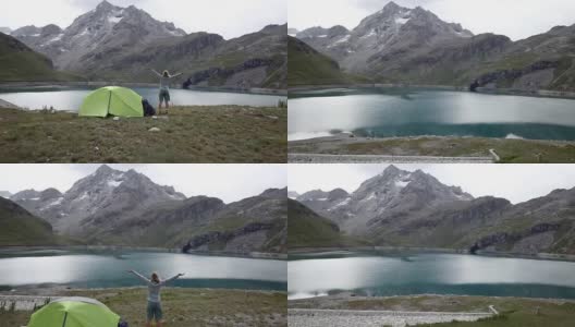 无人机拍摄，一名年轻女子在徒步旅行站在她的帐篷附近美丽的山湖双臂伸展高清在线视频素材下载