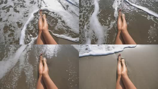 海浪冲刷着晒黑的女性脚。在一个阳光明媚的日子里，一个美丽的女孩躺在沙滩上。暑假期间，一个陌生的年轻女子在海边放松。俯视图慢动作特写高清在线视频素材下载