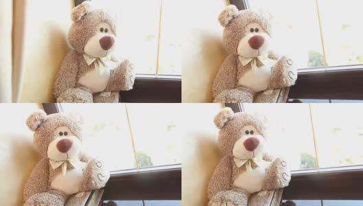 泰迪熊坐在窗台上，可爱的泰迪熊靠近窗户，在她的脖子上有一只蝴蝶，浅棕色的，儿童的软玩具，一个泰迪熊在她的脖子上有一只蝴蝶高清在线视频素材下载