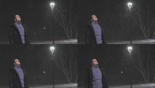 路灯下的那个人。在灯光的映衬下，正在下雪。慢动作，每秒180帧。雪花很漂亮。那个男人穿着一件凉爽的黑外套。高清在线视频素材下载