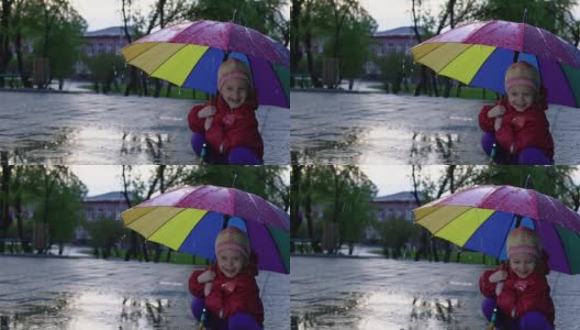 可爱的小女孩拿着五颜六色的雨伞在夕阳下的公园里的水坑里跳跃。高清在线视频素材下载