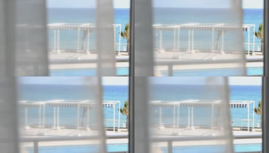 手持效果的镜头从一个人站在窗前看蓝色的大海或海洋透过飘动的透明白色窗帘在阳光明媚的夏天高清在线视频素材下载