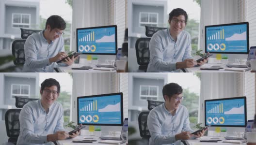 肖像头的年轻有吸引力的亚洲男子坐在微笑工作的多屏幕电脑和智能平板电脑在桌子上在家里的概念自由职业者数据分析师，数据科学家的业务。高清在线视频素材下载