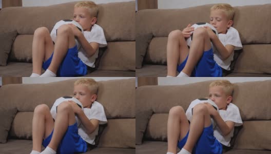 一个小男孩正坐在家里的沙发上，手里拿着一个足球。高清在线视频素材下载
