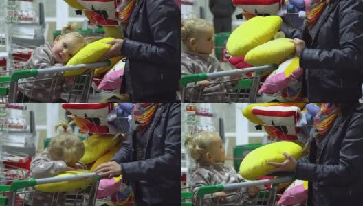 一位带着孩子的年轻妇女在超市选购商品。妈妈正用手推车载着一个小女孩。妈妈和女儿在看枕头。高清在线视频素材下载