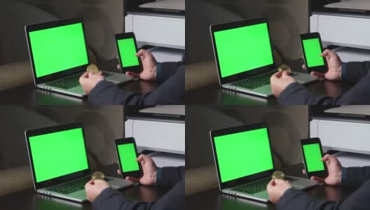 一名男子在一台绿色屏幕的笔记本电脑前工作。他手里拿着一部绿色屏幕的智能手机。考虑加密货币。Chromakey。高清在线视频素材下载