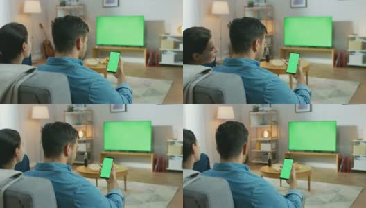 一对幸福的夫妇坐在客厅里看绿色屏幕电视，在沙发上放松。Guy还使用绿色模拟屏幕智能手机。高清在线视频素材下载