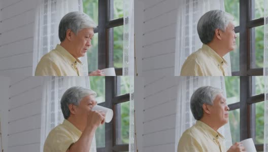 年长的亚洲男人站在窗边喝咖啡，放松情绪，退休生活高清在线视频素材下载