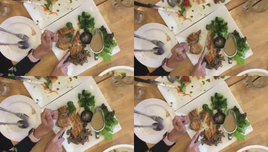 女人手拿刀叉切烤牛排在白色的盘子和食物，另一个是完成在餐馆的木桌上。前视图。视频片段。高清在线视频素材下载