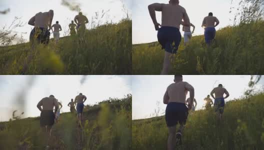 一群年轻人在蓝天上跑上绿色的小山，太阳耀斑在后面。日落时分，男运动员在大自然中慢跑。日出时在户外爬山的跑步者。慢动作高清在线视频素材下载