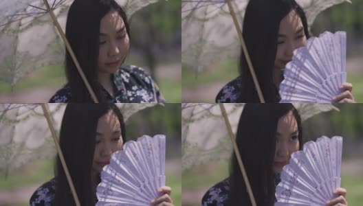 特写迷人害羞的亚洲年轻女子拿着白色太阳伞摇扇微笑。美丽的日本千禧一代穿着和服，在春天的户外公园享受阳光。高清在线视频素材下载