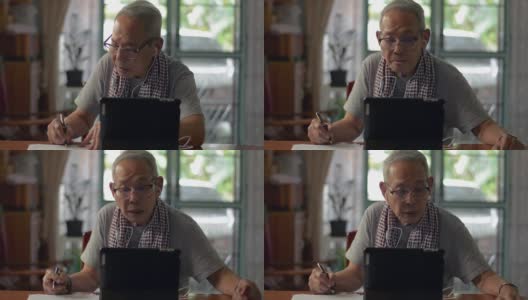 一位亚洲老爷爷穿着便装，在家用电子平板电脑上观看在线学习视频教程，并在纸上写字。老年人教育技术。高清在线视频素材下载