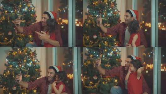 父亲和女儿与他们的家人视频通话，靠近圣诞树和客厅墙上的玻璃在圣诞节，4K分辨率。高清在线视频素材下载