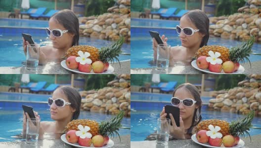 年轻美丽的女人用手机在游泳池旁边的水和盘子与热带水果在慢动作。hd 1920 x1080高清在线视频素材下载