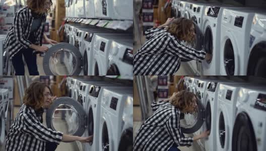 一个穿着格子衬衫的年轻女人正在家电商店里挑选洗衣机。打开门往里看。侧视图。慢动作高清在线视频素材下载