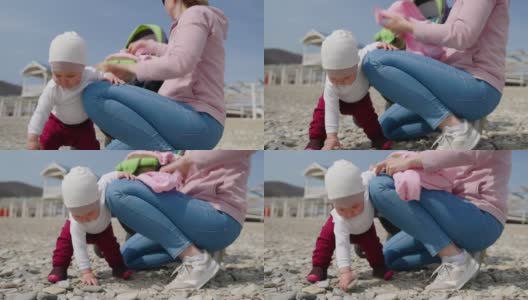 一个可爱的蹒跚学步的孩子捡起鹅卵石并仔细检查，而她的妈妈正在给她换衣服。儿童节的概念。实时高清在线视频素材下载