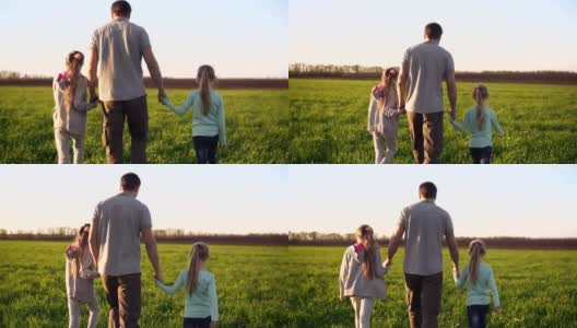 一个强壮的，年轻的父亲和两个小女儿走在绿色的草地上的背影。双手从两边握住它们。春季的一天。跟踪录像高清在线视频素材下载