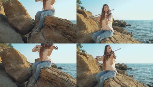 年轻漂亮的白人女孩穿着牛仔裤在美丽的海边拉小提琴，女人坐在大岩石上。慢动作高清在线视频素材下载