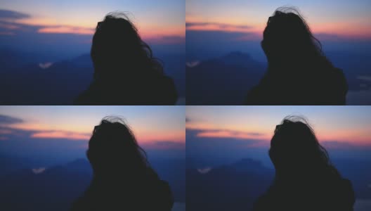 长棕色头发的徒步旅行者女孩在视点看日落在蓝色小时。风吹女人的头发在高山风景和湖泊。60帧/秒的慢镜头山峰高清在线视频素材下载
