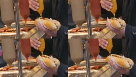 热狗。街道垃圾食品。男性的手拿着香肠包，把它和芥末倒在特殊的容器里高清在线视频素材下载