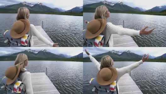 年轻女子站在美丽的山湖的木制防波堤上，双臂伸向自由。高清在线视频素材下载