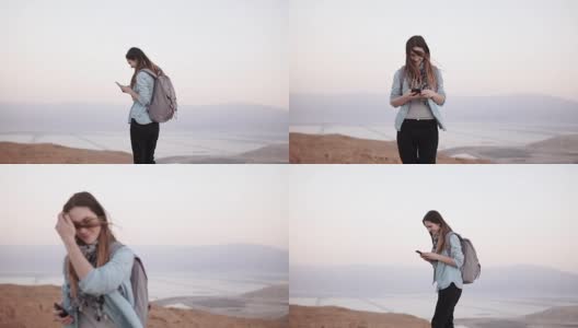 黄昏时分，一名女子在使用智能手机欣赏山景。缓慢的运动。messenger应用中背包类型的休闲女孩。以色列高清在线视频素材下载